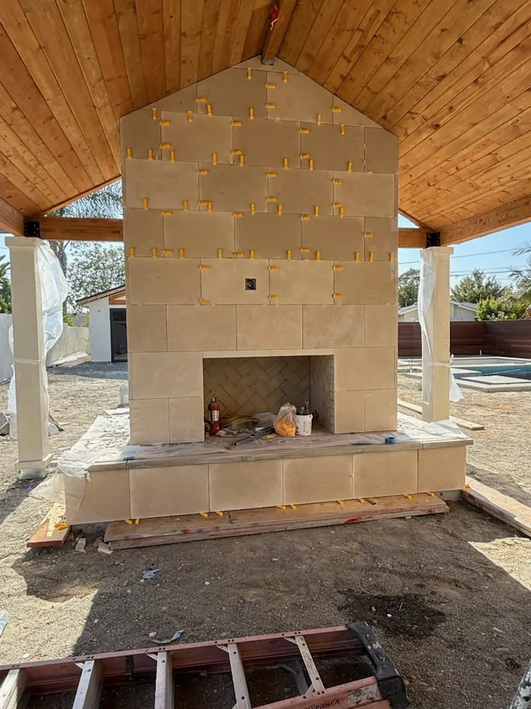 Outdoor stone fireplace under a wood pergola with a backyard pool in view